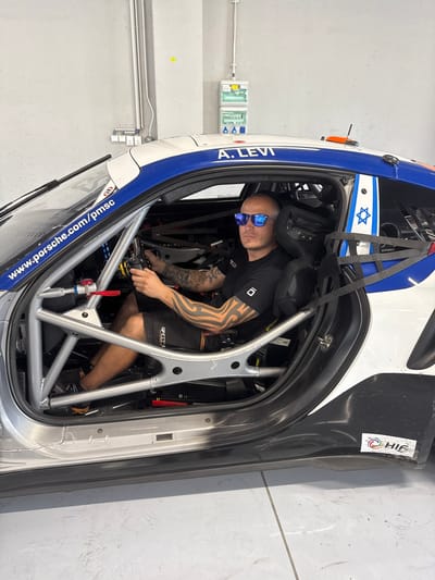 A man wearing sunglasses and a black t-shirt with the number "10" is seated inside a race car. The car's frame is visible, along with roll cage bars. The car features a blue and white design with the name "A. LEVI" printed on the side, near a star of David symbol. A website address is also visible on the car's exterior. The man appears to be adjusting something inside the car. A light green emergency sign is mounted on the wall behind. The background shows a clean, industrial space with a concrete floor.