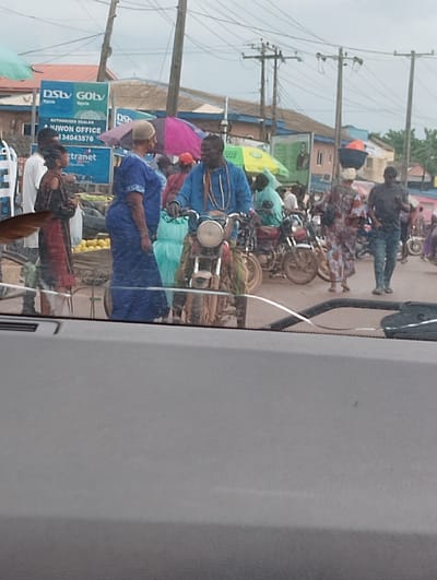 The image shows a busy street scene in Aketu-Oja, Nigeria. Several people are present, some standing and others on motorcycles. A man in a blue jacket is seated on a motorcycle, talking to a woman in a blue dress. Another woman, in a dark dress with red accents, stands nearby. In the background, a sign advertising DStv and GOtv is visible. Several motorcycles are parked or moving on the street. The environment appears to be an outdoor market or roadside area with various stalls and shops. Overhead power lines and utility poles are also present. The ground is a dirt or paved road. The sky is overcast.