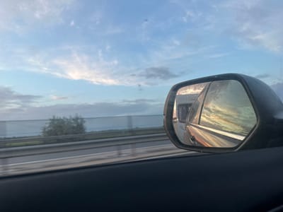 This image captures a scene from inside a vehicle, looking out at the road and sky near Óföldeák, Hungary. The car's side mirror, positioned on the left side of the frame (assuming standard viewing orientation), reflects a vibrant sky bathed in golden, orange, and reddish hues, indicative of either a sunrise or sunset. Within this reflection, part of a large vehicle, possibly a truck or RV, is visible behind, with some blurred, reversed text that is difficult to discern clearly, but suggests "MOTOR OIL" in reverse.
Through the car's side window, visible beyond the mirror, a multi-lane road or highway structure stretches horizontally across the upper portion of the scene. Below this road, the outline of a green tree can be seen in the middle distance. The background is dominated by a clear blue sky interspersed with fluffy white and grey clouds. The warm, soft light suggests the time of day is either early morning or late afternoon, as the sun is low on the horizon. The image conveys the feeling of movement and travel.