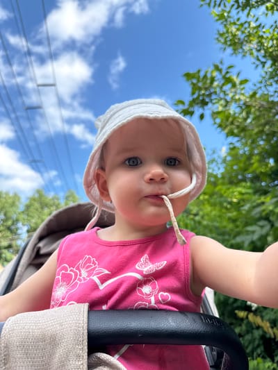 This is a close-up, vertically oriented photograph of a baby wearing a white sun hat and a pink tank top with a floral design. The baby is looking directly at the camera with a neutral expression. A string from the sun hat is in the baby's mouth. The background consists of a bright blue sky with scattered white clouds and blurred green foliage, suggesting an outdoor setting on a sunny day. A black stroller handle is visible on the right side of the frame. The image appears to be taken from a low angle, looking up at the baby. There is no visible text in the image. The scene is likely taking place during the daytime.