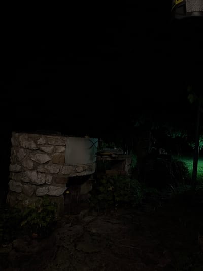 In the city of Chukovezer, Bulgaria, at night, a stone structure resembling a well or an old oven stands prominently. The structure is built with rough, irregular stones and has a metal panel on its front. To the right of the stone structure, a patch of green grass is illuminated by a light source, suggesting an outdoor setting with vegetation and possibly a yard or garden area. The overall scene is dark, with the exception of the illuminated grass and some faint lighting on the stone structure itself, indicating the absence of daylight. The image captures a quiet and possibly secluded outdoor space.
