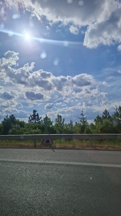 The image depicts a roadside view under a bright, partly cloudy sky with the sun shining strongly in the upper left quadrant, creating a lens flare. A guardrail runs horizontally across the middle of the frame, separating the asphalt road in the foreground from a dense line of trees and vegetation behind it. A triangular road sign with a graphic of a tunnel is visible behind the guardrail, to the left of center. The road surface is asphalt with a white line marking. The environment suggests a rural or suburban setting, possibly along a highway outside of a town. The location is specified as Malo Buchino, Bulgaria. There are no people or animals visible in the image.