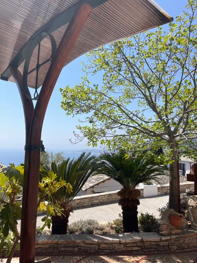 The image shows a view from a patio overlooking the Aegean Sea. The patio is located in the village of Moni Archaggelou, Greece at coordinates (40.597, 24.701).  A wooden pergola with slatted roofing shades a stone wall and a stone patio.  The stone wall encloses the patio and extends towards the sea.  Two palm trees stand in front of the wall, one on the patio and one on the hillside.  A third palm tree stands on the patio near the stone wall.  The sky is blue and clear with a few clouds.  In the background, a low white building with a red roof is visible.  There are no people in the image.