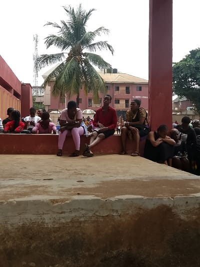 The scene depicts an outdoor area in Umuokpu, Nigeria. In the foreground, a concrete platform exhibits dirt and weathering. Directly behind this, a reddish-brown concrete ledge is visible. A group of approximately 15 individuals, primarily young adults, occupies the midground. A woman wearing a light pink top and leggings is seated on the ledge, looking down at a mobile phone held in her hands. Adjacent to her on the right, a man in a red collared shirt and dark shorts sits on the ledge, facing forward with legs crossed at the ankles. Further right, another woman in a dark patterned top is seated on the ledge. A large reddish-brown concrete pillar obstructs the view of additional individuals positioned further right and below the ledge, one of whom is a woman in a dark dress seated with her knees drawn to her chest. To the left, a reddish-brown wall is partially visible. In the background, a tall palm tree with coconuts stands prominently. Behind the tree, a three-story reddish-brown building is identifiable with "ST. JOSEPH SCHOOLS" inscribed on its facade. A communications tower is visible to the far left in the background. The sky is overcast.