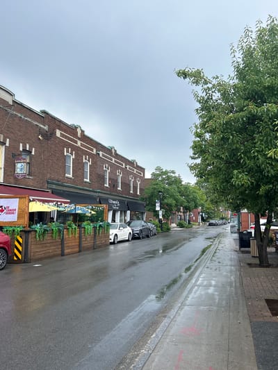 The image depicts a street scene in Sainte-Anne-de-Bellevue, Canada, on a cloudy day with wet pavement, suggesting recent rain. The street is lined with brick buildings on the left, featuring storefronts with outdoor seating areas covered by umbrellas and awnings. One storefront has a sign that says "GO CANADA GOOAL!". A red car is partially visible on the left, and several white and dark-colored cars are parked along the street. The right side of the street has a sidewalk and a tree. The sky is overcast and a light gray.