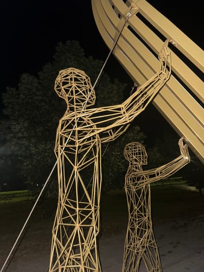 The image captures a nighttime scene in Egorlykskaya, Russia, featuring two wireframe sculptures of human figures. The figures are constructed from thin, golden-colored metal rods, forming a skeletal representation of a person. Both figures appear to be reaching upwards, with their arms extended towards a large, curved, golden structure overhead, which is likely part of the same art installation or a canopy.
The foreground sculpture is closer to the viewer, allowing for a detailed view of its geometric construction. Its head is a spherical cage of rods, and its body is a complex network of triangles and squares. The second sculpture, positioned further back and to the right, is partially obscured and appears smaller due to its distance.
The background is dark, indicating it is nighttime. Silhouetted trees with dense foliage are visible behind the sculptures, suggesting an outdoor setting, possibly a park or public space. The ground appears to be paved or a dark, wet surface, reflecting some light. A thin white cable runs diagonally across the lower portion of the image, adding a linear element to the composition.
The lighting in the scene seems to come from artificial sources, illuminating the golden sculptures and casting faint reflections. The overall mood is somewhat abstract and artistic, with the geometric forms of the sculptures contrasting against the organic shapes of the trees and the darkness of the night. There is no visible text in the image.