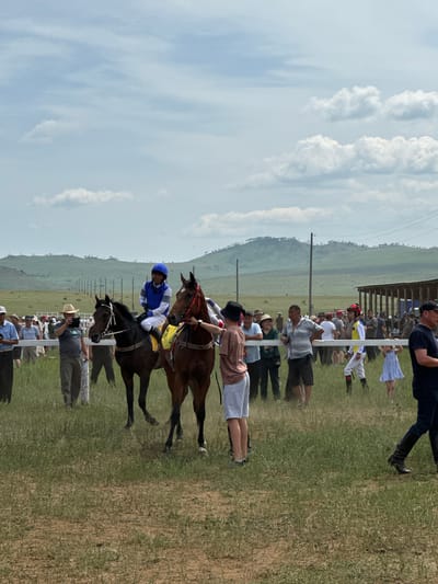 The image depicts a horse race event in Orongoi, Russia. The scene features several people gathered on a grassy field, with two horses and their jockeys in the foreground. One jockey, wearing a blue and white uniform, is mounted on a dark brown horse with a yellow saddle pad. Another jockey on a similar horse is partially visible to the right. Spectators, dressed in casual attire, are standing behind a white fence. In the background, a wooden structure, likely a stable or grandstand, is visible, along with rolling hills and a line of utility poles. The sky is partly cloudy.