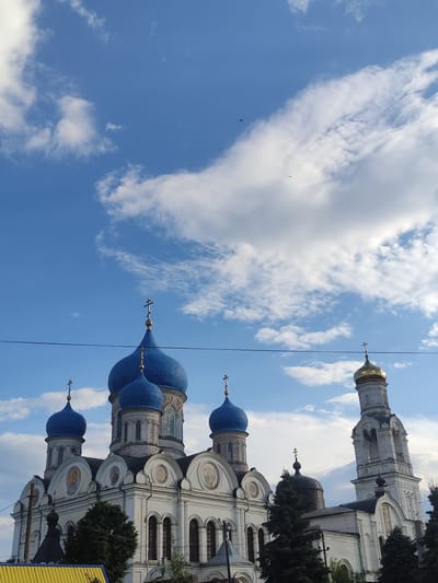 The image shows an outdoor view of the Ascension Cathedral in Rogachevo, Russia. The cathedral features multiple blue domes, one of which is capped with a golden cross, and a tall white bell tower with a golden dome. The building is predominantly white with ornate architectural details. Several evergreen trees are visible in front of the cathedral, and a bright yellow structure is partially visible in the lower left corner. The sky above is a vibrant blue, dotted with fluffy white clouds. A thin dark wire stretches horizontally across the upper portion of the image, and a few small birds are captured in flight against the sky. The scene is lit by natural daylight, suggesting it was taken during the day.