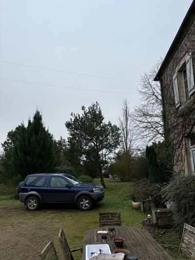 The image, rotated 90 degrees clockwise, presents an outdoor scene of a garden or backyard under an overcast sky. A dark blue, older model SUV, resembling a Land Rover Freelander, is parked on a grassy area in the mid-ground, facing left. The vehicle has silver alloy wheels and appears to be in a residential setting.

To the right of the parked car, a rustic wooden outdoor dining set, consisting of a table and chairs, is positioned on a patch of earth or gravel. On the table, several terracotta pots or decorative items are visible, along with a white rectangular plastic container holding various small objects.

The background features a variety of trees and bushes. Some deciduous trees are bare, suggesting late autumn or winter, while evergreen trees, including a prominent conifer, retain their foliage. A two-story building, likely a house, is partially visible in the top left corner, covered in climbing vines. It features windows with white shutters, indicative of European architecture. The ground is primarily green grass with some earthy patches. Thin power lines traverse the uniform grey sky above. The scene is devoid of people and discernible activity, portraying a quiet, static moment in Étréchy, France. No visible text is present.