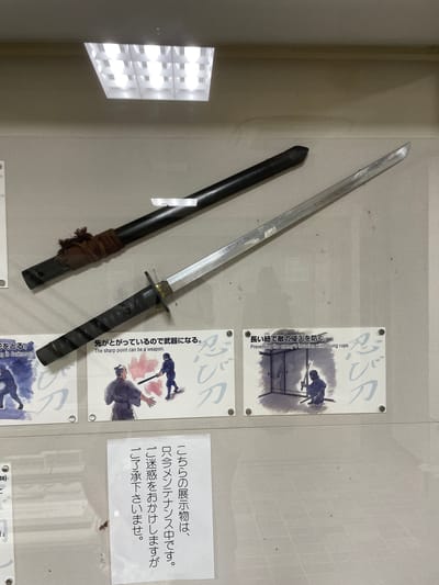 In Iga, Japan, this image captures a display featuring two Japanese swords, likely ninja swords (ninjatō), presented on a textured wall. The swords are positioned diagonally, with the lower sword, the ninjatō itself, extending from the bottom left to the top right of the frame. It has a dark, wrapped handle and a gleaming silver blade. Above and slightly behind it, another darker, cylindrical object, possibly a scabbard or another type of sword, is visible, with a brown fabric wrap near its hilt.

Below the swords, three illustrative panels are displayed. The leftmost panel depicts a stylized illustration of figures in motion, with the text "ing in darkness." to its left. The central panel shows two figures, one in traditional Japanese attire and another in what appears to be ninja garb, engaged in combat. The text above this panel reads, "The sharp point can be a weapon." The rightmost panel illustrates a ninja crouching indoors with a sword, and the text above it states, "Preventing the enemy's invasion with a long rope."

A separate, white informational sign is placed in the lower right quadrant of the image. It contains Japanese text, likely conveying information about the exhibit or maintenance. The background of the scene is a neutral, textured wall, and a modern fluorescent light fixture is visible on the ceiling in the upper portion of the image. The overall impression is of an educational exhibit about ninja artifacts or history.
