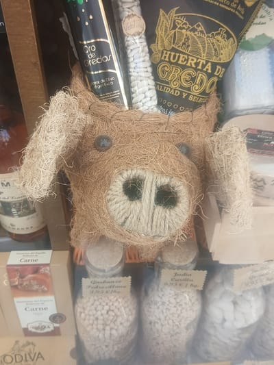This is a close-up, slightly angled shot of a display of food products, likely in a shop or market. The focal point is a whimsical teddy bear-like figure made from coarse, straw-like material, with prominent rope detailing for its nose and eyes. The bear is positioned centrally and appears to be lying down.
Surrounding the bear are various food items packaged in bags and bottles. Visible text indicates some of these are "Judia Carilla" (beans) priced at 6.95 €/kg and "Garbanza Pedrosillano" (chickpeas) at 3.95 €/kg. There is also a bottle of "Oro de Gredos Aceite de Oliva Virgen Extra" (Gredos Gold Extra Virgin Olive Oil). Further items include packages labeled "Pimientos del Piquillo Rellenos de Carne" (Piquillo peppers stuffed with meat) and "GODIVA". Some labels mention "Huerta de Gredos" and "Ávila", suggesting a Spanish origin, likely from the province of Ávila.
The lighting is somewhat diffused, and the overall impression is of a curated display of artisanal food products with a charming, handmade element. The scene appears to be indoors, given the lack of natural light and the organized presentation. There are no people visible in the frame. The overall mood is rustic and inviting.