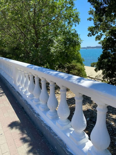 A low-angle, close-up shot shows a white balustrade extending along a paved walkway under a clear blue sky. The balustrade has a series of decorative, bulbous balusters. To the right of the balustrade, a sandy beach slopes down to the blue sea, with a distant coastline featuring buildings. Lush green trees with dense foliage frame the scene on the left and right, casting dappled shadows on the walkway. The environment appears to be a scenic overlook or promenade in Vinogradnoe, Russia, likely near the Black Sea.
