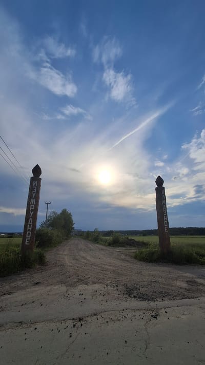 The image depicts a rural or semi-rural landscape located in Oliki, Russia, centered on a unpaved dirt road. The scene is illuminated by a bright, low-angled sun, positioned slightly left of center in the sky, partially obscured by thin clouds. The sky above is predominantly blue with scattered white, wispy clouds. A distinct white contrail extends diagonally from the upper left to the upper right of the frame.
In the foreground, a rough dirt road, composed of packed earth and small stones, leads into the distance. The road surface exhibits tire tracks and areas of darker gravel or coarse aggregate. Flanking the immediate entrance to the road are two tall, dark-stained wooden posts, each topped with a pointed cap.
The post on the left features vertically oriented Cyrillic text, "ИЗУМРУДНОЕ" (IZUMRUDNOE), translating to "Emerald." Behind this post, utility poles with power lines are visible, extending along the left side of the road. Green shrubs and low vegetation line the immediate roadside. In the distant background on the left, cultivated fields and a faint structure are discernable.
The post on the right features vertically oriented Cyrillic text, "АЛМАЗНОЕ" (ALMAZNOE), translating to "Diamond." To the right of the road, green fields extend towards a distant line of trees or forest on the horizon. Green shrubs and grass also line the right side of the road.
The dirt road recedes into the middle ground, appearing to narrow and potentially curve as it approaches the distant tree line. No individuals are present in the image.