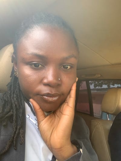 An individual with dark skin, dark dreadlocks, and a silver nostril stud is depicted in a close-up, chest-up shot within a vehicle in Nassarawo, Nigeria. The subject wears a dark grey pinstripe blazer over a white shirt, adorned with a necklace featuring large, clear, geometric stones. A gold-colored hoop earring is visible in the left ear. The subject's right hand is raised, resting with fingers splayed against the right cheek and jawline. The vehicle's beige interior headliner and seat are visible, along with a blurred outdoor scene through a rear window, suggesting bright ambient light illuminating distant vegetation and structures.