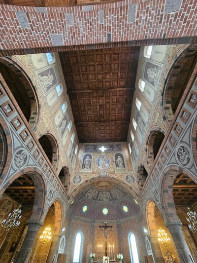 The image shows the interior of the Church of the Assumption of the Blessed Virgin Mary in Lentvaris, Lithuania. The photograph is taken from a low angle, looking up at the ceiling and the architecture of the church.

The ceiling is made of dark wooden planks, arranged in a coffered pattern. Supporting the ceiling are massive brick arches and columns. The walls are adorned with numerous fresco paintings, depicting biblical scenes and figures. These frescoes are in muted tones of beige, brown, and red, with some highlights of blue and yellow in the windows.

Along the nave, on the upper walls, are a series of arched windows letting in natural light. Below the windows, the walls are decorated with bands of intricate patterns and framed artwork, likely further religious imagery. In the foreground, pillars with decorative stone bases are visible, leading the eye towards the altar area.

The altar itself is not fully visible, but a cross with a figure of Christ is prominent, flanked by several lit candles and a small bouquet of white flowers. Chandeliers hang from the ceiling, providing artificial light, and some have an ornate, multi-tiered design. The overall impression is one of grandeur and historical significance, characteristic of old European churches. The architecture suggests a Romanesque or Gothic influence with the arched openings and stone construction.