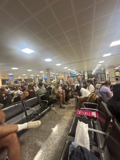 The image shows a crowded waiting area at Malta International Airport in Luqa, Malta. Passengers are seated on rows of black, airport-style seating, waiting for their flights. The scene is brightly lit by overhead fluorescent lights recessed into a white, grid-patterned ceiling. People of various ages and ethnicities are present, some looking at their phones, others resting. Luggage, including backpacks and bags, is placed on the floor or on the seats next to the passengers. In the foreground, a person's legs are visible with a white sneaker and white socks. Further down the waiting area, ATM machines and arrival/departure boards are visible.