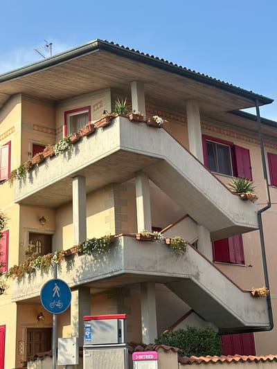 This is a medium-angle outdoor shot, oriented vertically, of a building's facade with a clear blue sky above. The architecture features prominent concrete staircases that zigzag up the building, adorned with rows of planters containing white flowers and some greenery. The building itself is painted in warm, muted tones of yellow and cream, with accents of dark red for the window shutters and frames. The roof has a dark, textured tile design.
In the lower right quadrant, a blue circular sign with a white pictogram indicates a shared path for pedestrians and cyclists. Adjacent to this sign are street furniture elements, including what appears to be a mailbox or utility box and a sign with the word "INTRED" in pink.
The lighting suggests it is daytime, with bright sunlight casting distinct shadows, particularly from the planters and architectural elements onto the building's walls. The overall impression is one of a well-maintained residential or commercial building, possibly in a European town, given the architectural style and the street sign. No people are visible in the frame. The scene evokes a sense of peaceful urban life.