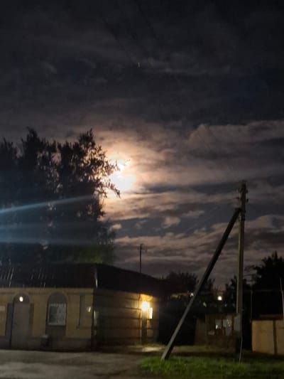 The image captures a nighttime scene in Novlyanka, Russia, illuminated by a bright full moon partially obscured by clouds. In the foreground, a low, single-story building with yellow walls and a dark roof is visible. The building has arched windows and doors, with some lights glowing from within. To the right of the building, a wooden utility pole stands at an angle, supporting wires that traverse the sky. Behind the building, silhouetted trees are visible against the moonlit sky. The ground in front of the building appears to be a paved or dirt surface, with a strip of grass and foliage in the lower right corner. The overall atmosphere is dark and somewhat eerie, typical of a rural or suburban night setting.