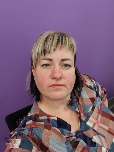 A medium shot, taken at chest level, captures a Caucasian woman with blonde and black hair, and blue eyes. She is seated in a black chair, facing forward, with her head tilted slightly to her left. Her hair is cut in a choppy style, with bangs covering her forehead. She is wearing a plaid shirt with red, blue, and white colors. The background is a solid purple wall. The image appears to be a selfie, taken indoors. The location is not specified in Prokudskoye, Russia.