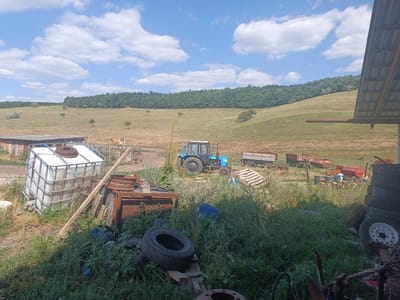 The image depicts a rural scene on a bright, sunny day with scattered white clouds in a blue sky. In the foreground, there is a collection of farm equipment and storage items, including a large white IBC tote with a metal cage, several rusty metal structures, a pile of tires, and overgrown green vegetation. A wooden beam leans against the IBC tote.
Further back, a blue tractor is parked in a grassy field. Behind the tractor, a row of agricultural trailers and equipment is visible. The landscape consists of gently rolling hills covered in dry grass, with a dense green forest line across the horizon. The overall impression is one of a working farm or agricultural storage area, likely located in a rural setting.
The image has been identified as being in Luminița, Moldova. However, there are no specific landmarks or contextual clues within the image itself that would definitively confirm this location. The scene is devoid of people.