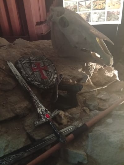 The image shows a still life arrangement of medieval-themed props, including a horse skull, a shield with a red cross, two swords, and an axe. The scene is set against a backdrop of rocks and sand. In the upper background, there is a display of multiple framed images, some appearing to depict historical or archaeological scenes. The lighting is dim, with a spotlight highlighting the central objects. The arrangement suggests a historical or fantasy exhibit, possibly within a museum or a themed attraction. The location is the Museum of the Earth in Sobótka, Poland, a place known for its geological and paleontological exhibits.