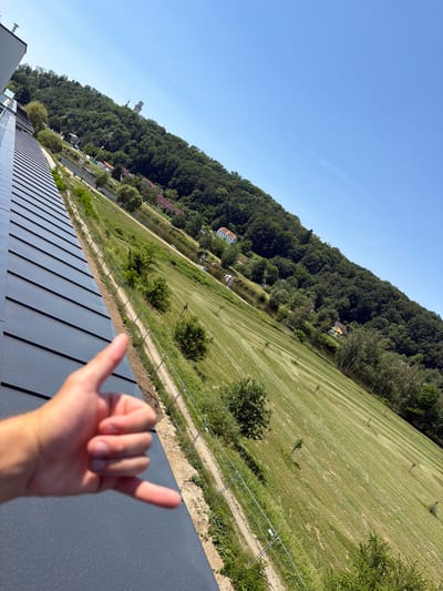 The image is taken from a high vantage point, looking down and across a sloped, green grassy field under a clear blue sky. In the foreground, a left hand is visible, with the thumb and index finger extended, forming a gesture resembling a "shaka" or "hang loose" sign. The hand is close to the camera and slightly out of focus.
The field is mown and shows distinct diagonal stripes from the mowing pattern. Scattered across the field are several small, young trees. A dirt path runs along the edge of the field, bordered by a metal fence. Beyond the path and fence, a body of water, likely a river or canal, is visible, with a few boats docked along its edge. On the opposite bank of the water, there are houses and more trees.
Dominating the background is a dense, tree-covered hillside that rises steeply. On top of the hill, a white tower structure is visible, along with what appears to be part of a castle or historical building complex. The overall scene depicts a sunny day in a landscape that combines urban elements (buildings on the hill) with natural features (forest, field, water).
Considering the described landscape and the presence of a notable tower/castle structure on a prominent hill overlooking a river, the location is consistent with Hluboká nad Vltavou, Czechia, specifically the area near the Hluboká Castle and its surrounding parkland, with the Vltava River in the vicinity. The modern building with solar panels in the lower left suggests a contemporary addition to the landscape, possibly a visitor center or modern facility.