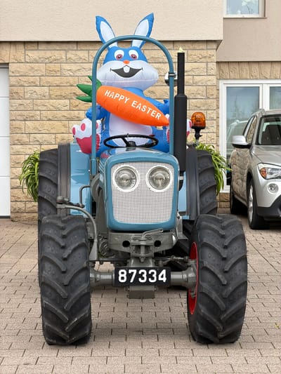 The image shows a blue tractor, with an inflatable Easter bunny attached to it, parked outside a building in Olingen, Luxembourg. The tractor has large, black tires and a white grill with two headlights. The license plate reads "87334". The building is made of beige brick, and a silver car is partially visible behind the tractor. The tractor is parked on a paved driveway.