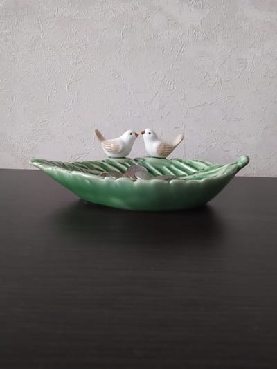 The image shows a decorative ceramic object shaped like a green leaf with two small white birds perched on it. One bird is positioned slightly above and behind the other. The ceramic leaf appears to be holding some coins within its curved form. The object is placed against a textured light-colored wall on the left and a dark wooden surface on the right. The lighting suggests an indoor setting, possibly a home. There is no visible text.