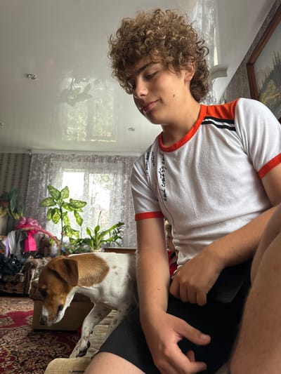 In an indoor setting, a young male with curly brown hair is seated on a couch, looking downwards. He is wearing a white t-shirt with orange trim and black detailing on the shoulders, and black shorts. His right hand rests on his lap, holding a dark object, possibly a phone. To his left, a medium-sized dog with white and brown fur stands on the couch, looking down and to the left. The background reveals a room with patterned wallpaper, sheer curtains covering a window, and several potted plants. A framed picture hangs on the wall to the right. The furniture includes a couch and an armchair, with a patterned rug on the floor. The overall impression is of a casual, domestic scene. The location is identified as Сароцкі сельскі Савет, Belarus.