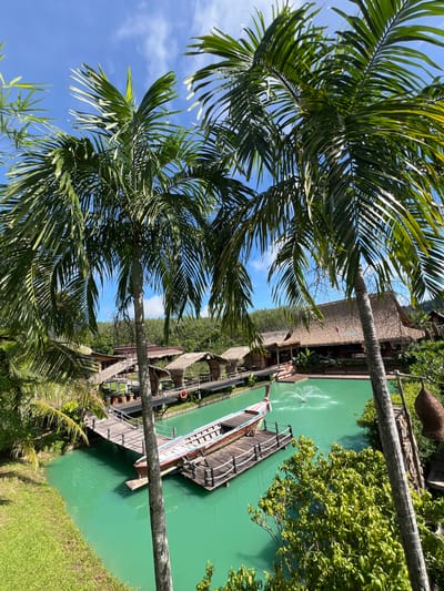 This is a photograph taken from an elevated vantage point in Tambon Pa Kae Bo Hin, Thailand. The scene is dominated by a vibrant turquoise body of water, likely a man-made pond or lagoon. On the water, a longtail boat, a traditional Thai vessel, is moored to a wooden pier. The pier extends from the shore, connecting to several thatched-roof structures nestled among lush greenery. These structures appear to be rustic huts or bungalows, suggestive of a resort or eco-tourism destination. In the background, more thatched-roof buildings are visible, possibly restaurants or communal areas, with their roofs blending into the dense foliage. Two large palm trees frame the foreground of the image, their fronds casting shadows and adding to the tropical ambiance. The sky is a clear blue with a few wispy white clouds. A fountain sprays water in the center of the pond, adding a dynamic element to the otherwise tranquil scene. The overall impression is one of a peaceful, tropical retreat.