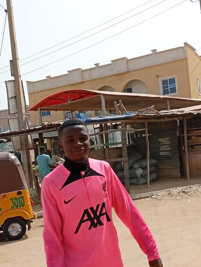The image captures a vibrant street scene in Fanisau, Nigeria, under bright daylight. The main subject is a young man, likely in his late teens or early twenties, positioned prominently in the foreground, slightly to the left of the center. He is looking directly towards the viewer with a subtle, pleasant smile or smirk.
He is wearing a distinctive bright pink long-sleeved athletic shirt, which features black panels on the upper chest and collar, a black zipper, and a black stand-up collar. Visible logos on his shirt include a black Nike swoosh on the right chest, the "LFC" (Liverpool Football Club) crest on the left chest, and the prominent "AXA" sponsorship logo in large black letters across the torso.
To the left of the man, the front portion of a yellow motorized tricycle, commonly known as a Keke Napep or auto-rickshaw, is partially visible. The number "3010" is clearly printed on its side. In the background, behind the man and the tricycle, are several makeshift market stalls or shelters constructed from wooden poles and topped with corrugated metal or fabric roofs. Within one of these stalls, several large, dark sacks are stacked. Another individual, dressed in a light blue garment, can be seen in the deeper background, near the market structures.
Further beyond the market stalls, a light yellow or beige two-story building with a flat roof is visible. It features arched windows on the lower level and rectangular windows with white frames on the upper level. The sky above is clear and bright, indicating a sunny day. The ground in the foreground and visible street areas appears to be unpaved and dusty. The overall scene suggests a bustling, everyday environment in an urban or semi-urban setting.