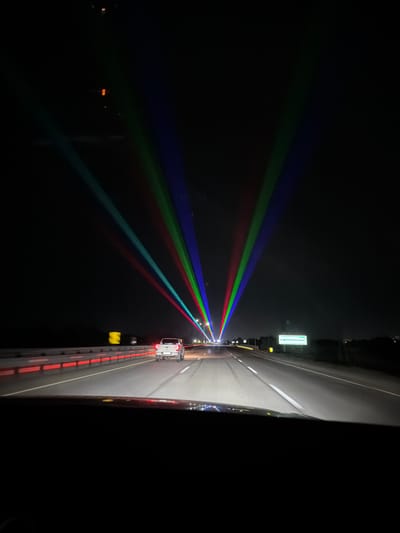 The image captures a nighttime view from inside a vehicle traveling on a highway near Zarechnoye, Kazakhstan. Multiple beams of colored light, predominantly red, green, and blue, are projected into the sky from an unseen source, creating a spectacle of light trails. The highway ahead is illuminated by the vehicle's headlights and the distant streetlights. A white pickup truck is visible in front, traveling in the same direction. The road is flanked by barriers with red lights and a green directional sign is visible on the right side of the road. The sky is dark, indicating it is nighttime, and the overall atmosphere suggests a potentially festive or event-driven lighting display.