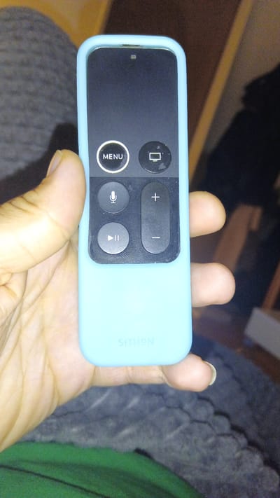 A close-up, slightly high-angle shot depicts a hand holding an Apple TV remote, encased in a light blue silicone cover. The hand, with a light-to-medium skin tone and natural-looking fingernails, cradles the device centrally in the frame.

The Apple TV remote features a prominent black, glossy surface for its buttons and touchpad, visible through an opening in the protective case. The top-left circular button displays the text "MENU," while the top-right button features a television screen icon. Below these, a circular button on the left shows a microphone icon, indicating voice control, and a vertical pill-shaped button on the right is marked with a "+" at the top and a "–" at the bottom, suggesting volume control. The bottom-left button is circular and displays a play/pause icon. A tiny light indicator is visible at the very top edge of the remote, above the black touchpad area.

The light blue silicone case has the brand name "SITHON" embossed near its bottom. The background is out of focus, showing a textured, dark grey surface, likely a blanket or clothing, on the left and bottom, with a hint of green fabric in the lower left corner. The right side of the background is darker and less defined. The overall lighting is dim, suggesting an indoor setting, possibly at night or in a softly lit room. No specific location cues for Ginsheim-Gustavsburg, Germany, are visible.