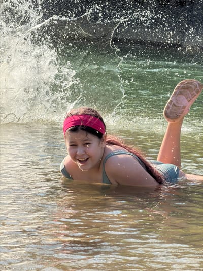 A young girl is lying on her back in water, with waves crashing around her. She is wearing a teal bikini top and a pink headband. Her hair is long and dark, and she is smiling broadly. Her right leg is extended out of the water, with a pink flip-flop on her foot. The water is a murky green, and the sunlight reflects off of it, creating a shimmering effect. There is a dark, rocky area to the left of the frame, where the waves are breaking and splashing. The scene appears to be taking place on a sunny day, likely at a beach or lake. The girl's expression conveys joy and excitement.