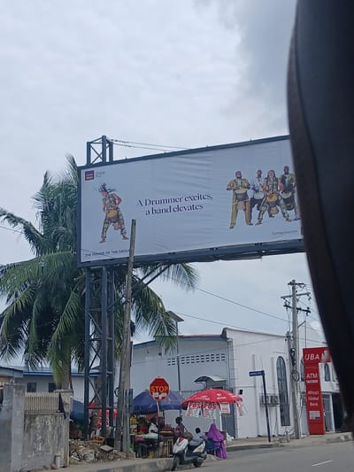 This media file shows an outdoor scene in Animashawun, Nigeria. The main subject is a large billboard featuring a group of drummers in traditional attire. The text on the billboard reads "A Drummer excites, a band elevates" and "fcmbgroup.com" with the FCMB Group PLC logo.
In the background, there is a UBA (United Bank for Africa) branch with prominent signage including "ATM" and "Africa's Global Bank". A street sign indicates "AMOSU STR". Several people are visible near a street-side market with stalls displaying fruits and other goods. An Airtel umbrella is also prominent. A red octagonal "STOP" sign is mounted on a pole. The sky is overcast, suggesting a cloudy day. The presence of palm trees indicates a tropical or sub-tropical climate. The overall scene depicts a busy urban environment with commercial activity and advertising. The image appears to be taken from a vehicle, as suggested by the partial view of the car's interior at the top of the frame.