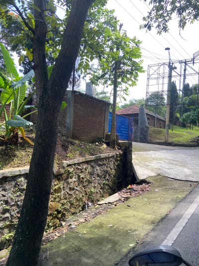 The image shows a street in Kecamatan Cisarua, Indonesia, located at latitude -6.84 and longitude 107.551. A large tree trunk is in the foreground, partially obscuring a stone wall and a drainage ditch. Behind the stone wall is a dirt road leading up a hill. On the left side of the road is a red brick building with a blue gate. A metal structure with a billboard, partially obscured by foliage, is visible on the upper right side of the image. The image is taken from a low angle, looking up the road. The image is clear, the colors are vivid, and the details are sharp. There are no people or vehicles in the image, but the presence of the motorcycle mirror suggests that the image was taken from a vehicle.