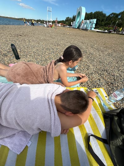 This image captures a bright, sunny day at a pebble beach in Taman, Russia, showing two children relaxing. The photo is taken from a high angle, looking down on the subjects.

In the foreground, two children are seen from behind, lying on towels on the stony shore. The child on the left, appearing to be a girl with dark hair tied back with a pink scrunchie, is partially covered by a brown patterned towel. She is wearing a swimsuit and seems to be looking towards the water and the background, with her hands resting on pebbles. The child on the right, appearing to be a boy with short dark hair, is covered by a light purple towel and is resting his head on his bent arm. He is lying on a yellow and white striped mat.

Next to the children on the mat, a clear plastic water bottle is visible, bearing the Russian text "СВЯТОЙ ИСТОЧНИК" (Svyatoy Istochnik, meaning "Holy Spring"). A dark green backpack or bag is also on the mat, with a black strap extending from it. Small, smooth pebbles are abundant across the entire beach surface.

In the mid-ground and background, the beach stretches along a calm, blue body of water. Large, light blue decorative letters, which appear to spell out "TAMAN," are erected on the beach, likely indicating the location. Behind these letters are lush green trees. Further down the beach, other people are visible, and a tall, white structure, possibly a lifeguard stand or observation tower, stands near the water's edge. The sky above is clear blue with a few scattered clouds. The strong sunlight casts clear shadows, suggesting it's either mid-morning or afternoon. The overall scene conveys a relaxed beach day.