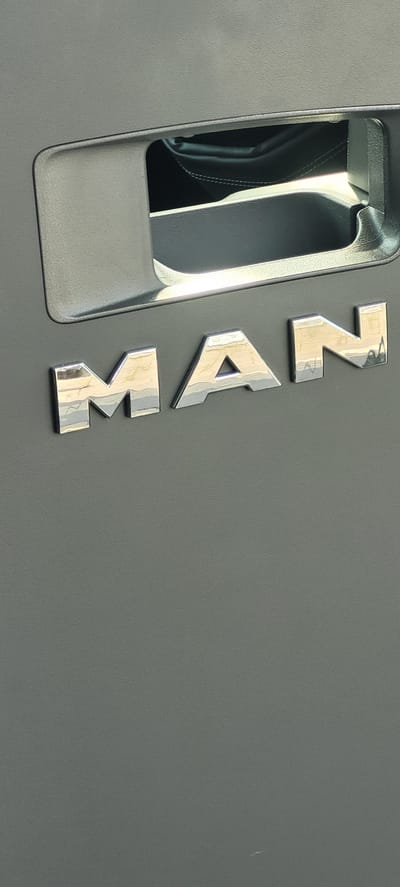 The image shows the word "MAN" in chrome lettering on a textured gray surface, likely the interior of a vehicle. Above the lettering, there is a recessed area with a dark interior, possibly a storage compartment or part of a door handle mechanism. The lighting suggests an indoor or shaded environment, with reflections visible on the chrome letters and the recessed area. There are no people visible in the image. The overall context of the image is a close-up of a vehicle's interior, specifically focusing on branding or labeling. The image does not contain any information that would allow for identification of its location in Obertshausen, Germany.
