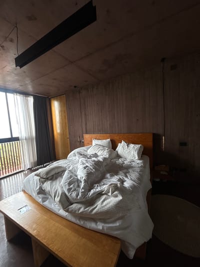 An interior view of a room in Tepoztlán, Mexico, reveals a minimalist design with a focus on natural light. The room features a large window with horizontal blinds that are partially open, revealing the outdoors. A double bed dominates the space, covered in white linens and several pillows. The bed frame is constructed from light-colored wood. The room's walls and ceiling are made of concrete and wood respectively, lending an industrial feel to the space. A prominent black steel beam runs along the wall. An area rug is visible, suggesting a defined seating or relaxation area. A wooden bedside table can be seen.
