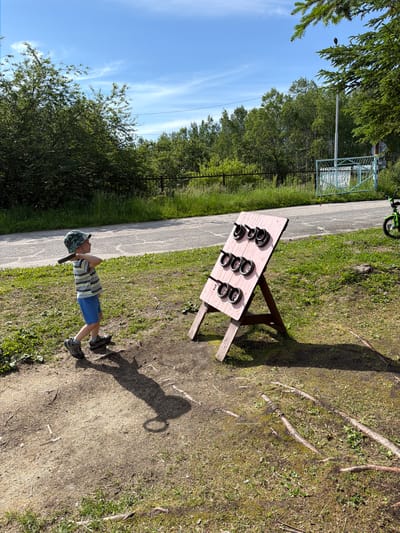 A young boy in a striped t-shirt, blue shorts, a patterned bucket hat, and grey sneakers stands on a dirt and grass patch, holding a dark object horizontally at shoulder height, appearing to mimic swinging a bat. He is facing towards a wooden game on an easel, which is painted pink and displays nine black rings in three rows of three. The game is set up on a sloped surface with grass and scattered twigs. In the background, a paved path is visible, bordered by more grass and a dark metal fence. Beyond the fence, lush green trees and foliage form a dense line, with a bright blue sky above featuring thin, wispy clouds. To the right of the game, a bright green bicycle with black tires is parked near a pale blue gate and a white structure. The scene is outdoors on a sunny day. The location is identified as being in the city of Kultdur, Russia.