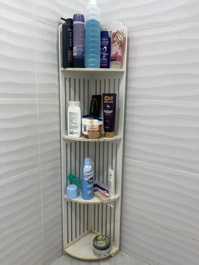 A white plastic shelf, divided into compartments, is attached to a white-tiled wall. The shelf holds various personal care products, including shampoos, conditioners, shaving cream, and hair masks. Product brands such as L'Oréal Elvive, Syoss, Chi Brilliance, and Balea Men are visible. One product label is written in Cyrillic: "Бадем Маска за коса" ("Badem Mask for Hair"). The curved right end of the shelf is positioned in a corner of the room, with more wall tiles visible. The specific location within the city of Kichevo, Bulgaria, cannot be determined from the image.
