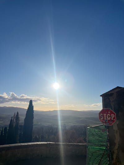 This image captures a bright, sun-drenched landscape under a clear blue sky, viewed from an elevated perspective, consistent with the Pienza region of Italy. The sun is a dominant feature, positioned in the upper-left, emitting intense rays and lens flare that extend horizontally across the frame.

The scene showcases vast, rolling hills and valleys characteristic of the Tuscan countryside, extending into a hazy distance. Scattered white clouds are visible along the horizon, illuminated by the bright sunlight. In the foreground on the right, a distinctive red octagonal "STOP" sign is attached to a stone building or structure. The sign has several small, illegible stickers or markings. Below it, a textured green screen or barrier is also visible.

Towards the bottom right, a tall, dark cypress tree stands prominently, with hints of other trees further back in the landscape. A stone or concrete wall forms part of the foreground at the bottom right, suggesting the vantage point is from a terrace or balcony. The image conveys a sense of serene, bright daylight, likely late morning or early afternoon. No people or specific activities are depicted, focusing instead on the expansive natural beauty and a few man-made elements. The only clear text visible is "STOP" on the red sign.