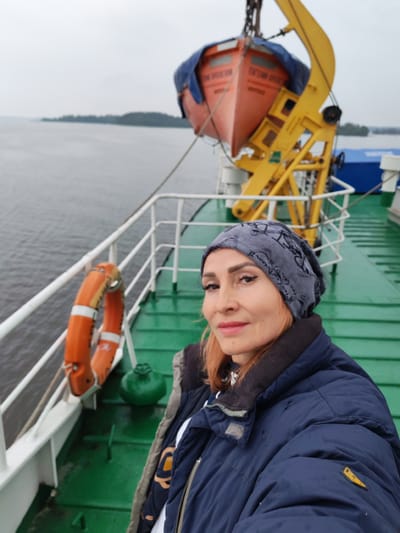An outdoor medium shot captures a woman from the chest up, facing forward with a slight smile. She wears a blue puffer jacket and a grey beanie with a patterned design. The background shows a green deck of a boat or ferry, a white railing, a life preserver, and a yellow crane holding an orange lifeboat. The water is visible to the left, with a distant, tree-covered shoreline under an overcast sky. The scene appears to be on the water in the vicinity of Goritsy, Russia, given the presence of a waterway and the characteristic landscape of the region.
