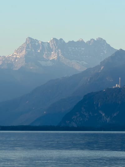 The image depicts a wide shot of a lake in front of layered mountains. In the foreground, the calm, blue water of a lake reflects the sky and surrounding mountains. The middle ground features a series of dark, tree-covered hills and mountains that recede into the distance, becoming increasingly hazy. A distinct structure, resembling a white tower and some buildings, is visible on a ridge in the middle distance, nestled within the dark, wooded slopes. The background is dominated by a range of imposing, rugged mountains, their peaks capped with snow and illuminated by soft light, suggesting either early morning or late afternoon. The sky above is a pale, clear blue, with no clouds visible. The overall impression is one of a serene, natural landscape with human intervention subtly present in the form of the tower and buildings. The scene is likely located in the vicinity of La Tour-de-Peilz, Switzerland, given the characteristic lake and mountain topography of the region.
