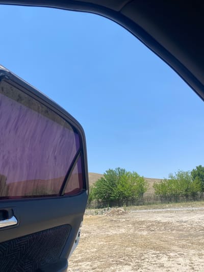 The image shows an open car door with purple-tinted windows, looking out onto a dry, dusty landscape under a clear blue sky. In the background, there are sparse trees and rolling hills characteristic of the region. This scene is consistent with the natural environment found near Naray, Kyrgyzstan, where such arid terrain and vegetation are common. There are no people visible in the image.