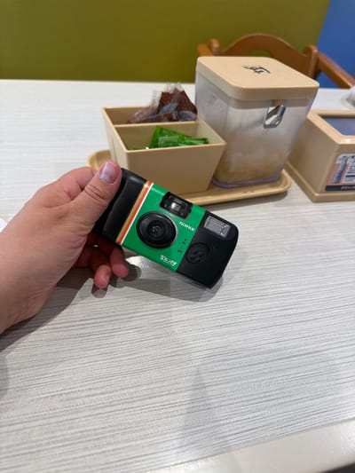 An over-the-shoulder medium shot shows a person's left hand holding a green and black Fujifilm disposable camera. The camera has white and gold stripes on its front. The person's hand is positioned on the right side of the frame, and their fingers are curled around the camera. The camera is oriented horizontally, with the lens facing away from the viewer. Behind the camera, a beige tray holds a small, white container with individually wrapped snacks. To the right of the tray, a clear plastic dispenser with a beige lid contains a light-colored substance. The scene takes place on a light gray, textured table. The background is blurred, but hints of a green wall and wooden chair can be seen. The overall setting appears to be a casual dining environment, potentially a cafe or restaurant within Chiyoda, Japan.