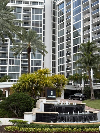 This image depicts the entrance to The Ritz-Carlton hotel in Bal Harbour Village, United States of America. A tiered water fountain with black tiled basins and white concrete accents is in the foreground, surrounded by meticulously manicured landscaping featuring various green shrubs and colorful low-lying plants. Behind the fountain, a dark-colored luxury SUV, possibly a Rolls-Royce, is parked on a paved driveway leading to the hotel.
The hotel itself is a towering, modern building with a white facade characterized by numerous floor-to-ceiling windows and balconies. Tall palm trees and other lush tropical foliage are interspersed around the building, adding to the resort-like atmosphere. A prominent sign for "THE RITZ-CARLTON" is visible to the left of the hotel entrance, adjacent to the fountain. The building's address, "10275", is also subtly displayed above the entrance. The overall scene conveys an image of luxury and upscale hospitality.