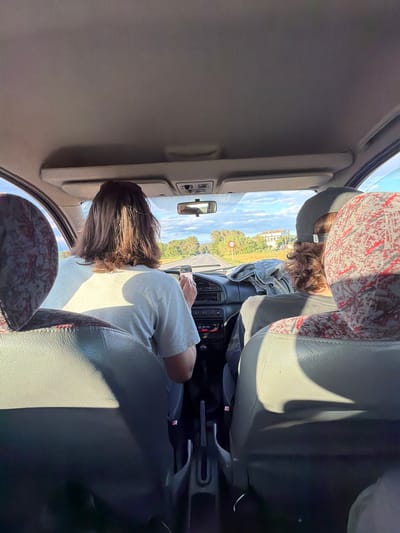 The image shows an interior car view. A person is driving while looking at a phone. Another person is in the back. Outside the car window, there is a road and green trees. The location is Torrent, Spain.