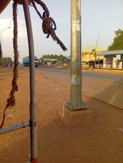 The image captures a street scene in Shuwarin, Nigeria, during what appears to be late afternoon or early morning, indicated by the warm light and subtle shadows. The foreground is partially obscured by a rustic, rope-like material and a metal pole structure. The ground is reddish-brown sand and dirt, with scattered dry vegetation and debris.
A paved road runs through the center of the image, extending into the distance. On the left side of the road, a blue truck is driving away, and further back, a person on a bicycle and a few pedestrians are visible near some small structures. On the right side of the road stands a large, galvanized metal utility pole with a robust concrete base. Attached to this pole are faded posters, one of which clearly shows two men's faces, likely political figures, with partially visible text "NNO" and "FAYAN...". Another poster or sign on the pole displays "GENERAL ENTERPRISE".
Behind the utility pole, a yellow building with a distinctive green-tiled roof and arched windows is prominent. Next to it are several smaller commercial establishments with metal roofs and gated fronts, one displaying "POS" (Point of Sale) on a sign. Further down the road on the left, another building shows text "LEMA DA POS". The sky above is clear and bright, with some trees visible in the background, completing the semi-urban or rural setting.