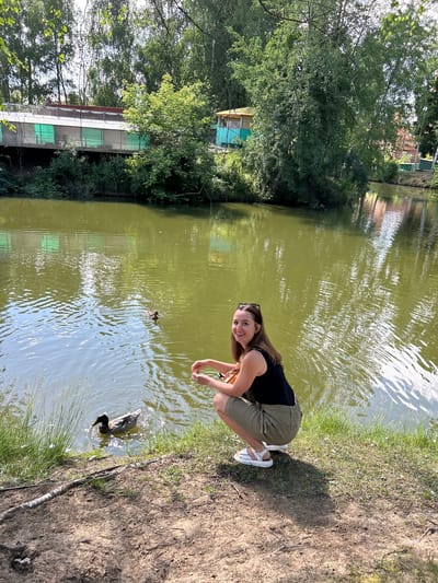 The image shows a young woman kneeling on the grassy bank of a pond, reaching out towards two ducks swimming in the murky green water. She is wearing a black tank top and olive green shorts, with white sandals. Her hair is brown and she is smiling at the camera. The pond is surrounded by trees and vegetation, and in the background, there are buildings that appear to be part of a park or recreational area. The sun is shining, casting a shadow of the woman on the ground, suggesting it is daytime. The overall scene evokes a peaceful and pleasant outdoor setting.